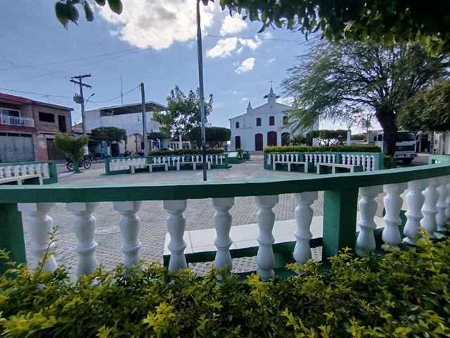 Vista da Praça Santa Terezinha em Cristais de Macaúbas, com árvores, bancos e moradores reunidos.
