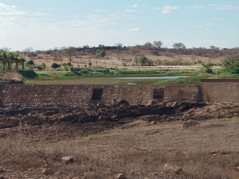 Leito seco do Rio Paramirim em Cristais de Macaúbas, com solo rachado, vegetação ao fundo e céu claro.